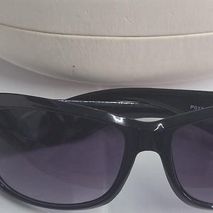 May include: Black sunglasses with dark tinted lenses and a thick, glossy frame. The sunglasses are positioned in front of a rounded, cream-colored hard-shell protective case. The frame features a smooth, continuous design with wide temples.