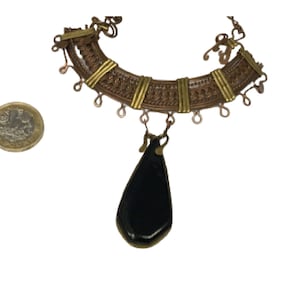Puede incluir: Un collar de oro y cobre con un colgante de lágrima negra. El collar tiene un diseño tejido y una serie de pequeños bucles que cuelgan de la parte inferior.