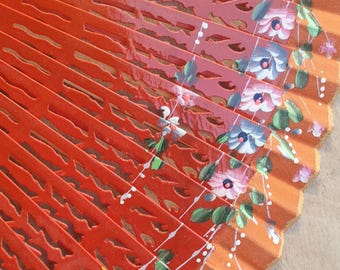 May include: A partially open, hand-painted fan with a vibrant red and orange color scheme. The fan features intricate cut-out designs and is adorned with delicate floral patterns in pink, white, and green. The fan's design is reminiscent of traditional Spanish fans.