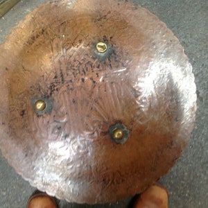 May include: A round copper tray with a decorative design and four brass accents. The tray has a hammered finish and a slightly raised edge.