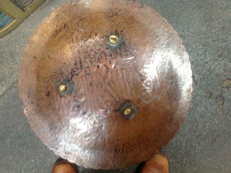 May include: A round copper tray with a decorative design and four brass accents. The tray has a hammered finish and a slightly raised edge.