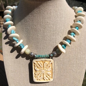 May include: A white and turquoise necklace with a carved bone pendant. The pendant has a square shape and intricate designs. The necklace is made with round white beads and turquoise stones.
