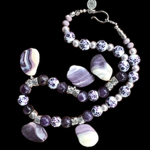 May include: A handcrafted necklace featuring a mix of purple and white beads, accented with silver-toned metal spacers. The necklace includes decorative purple and white shell-shaped pendants and a silver clasp. The beads have a floral pattern.