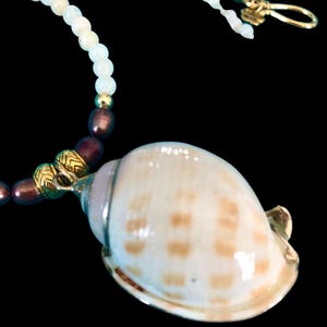 May include: A seashell pendant necklace with a cream-colored shell featuring brown spots. The necklace has a beaded strand of light green and dark brown beads, with gold-colored accents and a gold clasp.