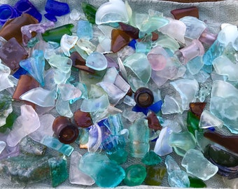 May include: A collection of sea glass in various colors and shapes, including blue, green, brown, and pink. The glass pieces are arranged on a white towel.