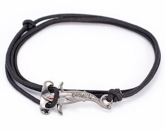 Könnte beinhalten: Schwarzes Kordelarmband mit einem silberfarbenen Hammerhai-Anhänger. Der Anhänger trägt die Gravur "Castaway". Das Armband ist mit verstellbaren Knoten gebunden.