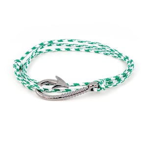 May include: A nautical-themed bracelet featuring a silver-colored fish hook clasp. The bracelet's cord is white with a green geometric pattern. The bracelet is designed to be worn around the wrist.