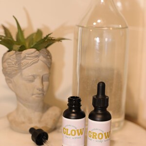 Puede incluir: Dos peque&ntilde;os frascos negros con goteros y etiquetas blancas. Las etiquetas tienen el texto "HerbBloom" y "Glow face serum" en una botella y "HerbBloom" y "Grow hair & scalp serum" en la otra. Los frascos est&aacute;n sobre una superficie de m&aacute;rmol blanco.