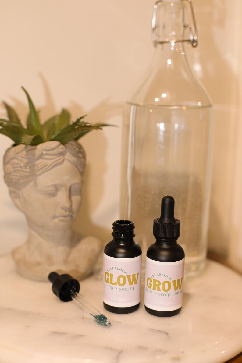 Puede incluir: Dos peque&ntilde;os frascos negros con goteros y etiquetas blancas. Las etiquetas tienen el texto "HerbBloom" y "Glow face serum" en una botella y "HerbBloom" y "Grow hair & scalp serum" en la otra. Los frascos est&aacute;n sobre una superficie de m&aacute;rmol blanco.
