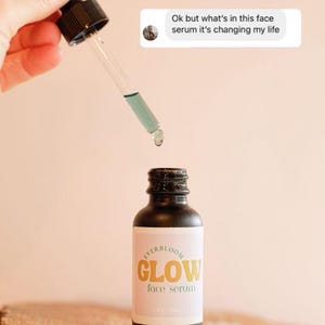 Puede incluir: Una botella de vidrio con un gotero negro y una etiqueta que dice "EVERBLOOM GLOW face serum". La botella est&aacute; sobre una superficie de madera. El gotero est&aacute; lleno de un l&iacute;quido azul claro.