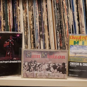 May include: Three vintage cassette tapes are displayed in front of a stack of vinyl records. The tapes feature album art for Richard Pryor's Greatest Hits, Sam Kinison's Leader of the Banned, and Cheech & Chong's Greatest Hit.
