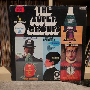May include: A black vinyl record album cover with the title "The Super Groups" in white lettering. The cover features illustrations of various artists and their hit songs, including The Rascals, Cream, Buffalo Springfield, Iron Butterfly, Bee Gees, Vanilla Fudge, and Mr. Soul.