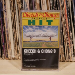 May include: A retro cassette tape of Cheech & Chong's Greatest Hit album. The cover art depicts a surreal scene with two figures supporting a large, cloud-like object. The cassette is in a clear plastic case.