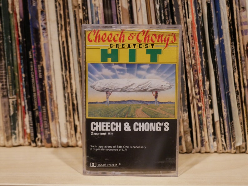May include: A retro cassette tape of Cheech & Chong's Greatest Hit album. The cover art depicts a surreal scene with two figures supporting a large, cloud-like object. The cassette is in a clear plastic case.