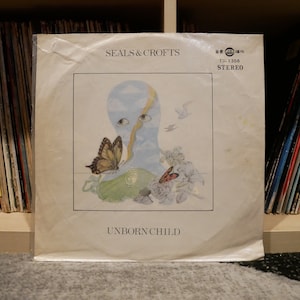 May include: Vinyl record cover for the album "Unborn Child" by Seals & Crofts. The cover features a blue and white illustration of a face with eyes and clouds. There are butterflies and flowers surrounding the face.