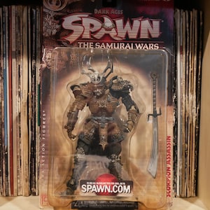 May include: A Spawn action figure from the Samurai Wars series. The figure is dressed in brown samurai armor with a horned helmet. The figure is holding a sword and is packaged in a plastic blister pack. The packaging features the Spawn logo and the text "The Samurai Wars".