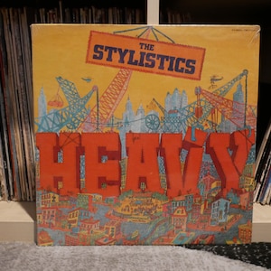 Op de afbeelding: Een vintage vinyl platenhoes met een kleurrijke illustratie van een stadsbeeld met kranen en de tekst "The Stylistics Heavy" in rode letters.