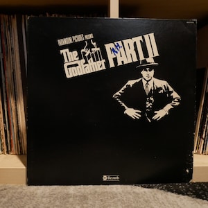 May include: A black vinyl record album cover for The Godfather Part II, featuring a black and white image of a man in a suit and tie, with the title text in white.