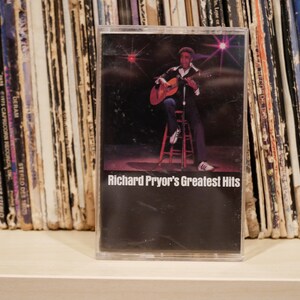 May include: A cassette tape of Richard Pryor's Greatest Hits. The cover shows Pryor playing a guitar. The cassette is in a clear plastic case, with a backdrop of record albums. The tape is vintage.