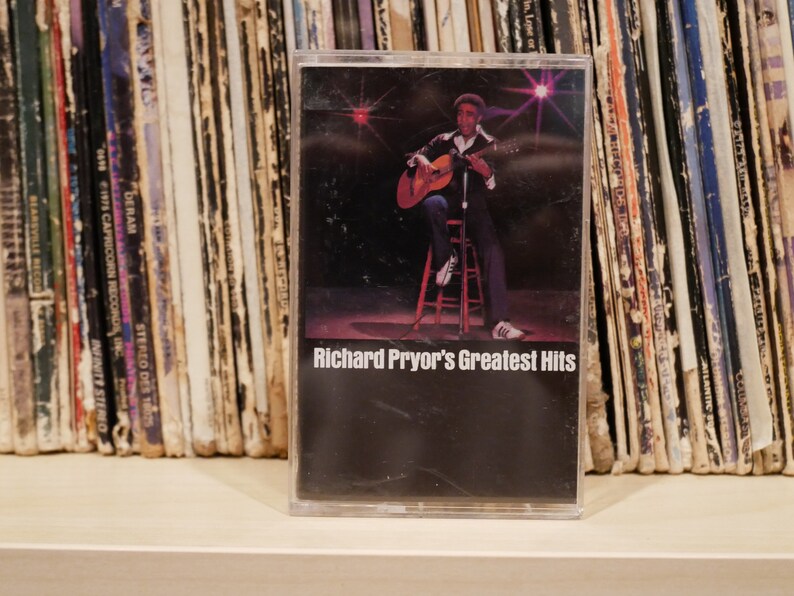 May include: A cassette tape of Richard Pryor's Greatest Hits. The cover shows Pryor playing a guitar. The cassette is in a clear plastic case, with a backdrop of record albums. The tape is vintage.