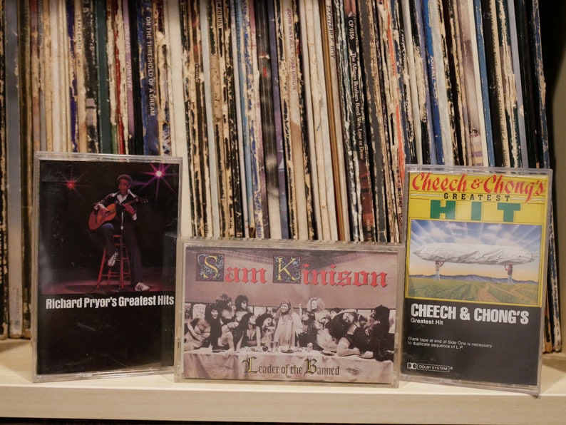 May include: Three vintage cassette tapes are displayed in front of a stack of vinyl records. The tapes feature album art for Richard Pryor's Greatest Hits, Sam Kinison's Leader of the Banned, and Cheech & Chong's Greatest Hit.