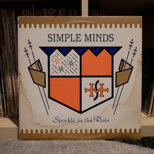 Peut inclure: Une pochette d'album de disque vinyle vintage avec un design graphique coloré avec un bouclier et le texte "SIMPLE MINDS Sparkle in the Rain".