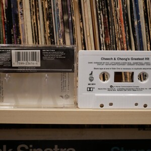 May include: A white cassette tape titled "Cheech & Chong's Greatest Hit" in a clear plastic case. The tape lists songs like "Earache My Eye" and "Basketball Jones." A collection of vinyl records forms the background.