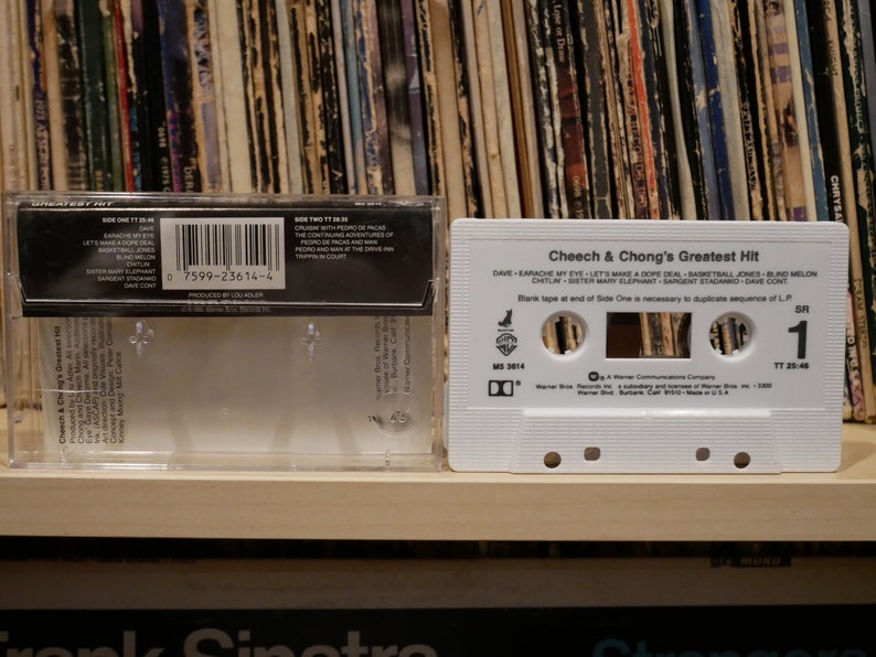 May include: A white cassette tape titled "Cheech & Chong's Greatest Hit" in a clear plastic case. The tape lists songs like "Earache My Eye" and "Basketball Jones." A collection of vinyl records forms the background.