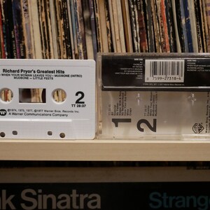 May include: A white cassette tape featuring "Richard Pryor's Greatest Hits" with song titles like "Cocaine" and "Mudbone." The cassette sits beside its clear plastic case, with a backdrop of vinyl records.