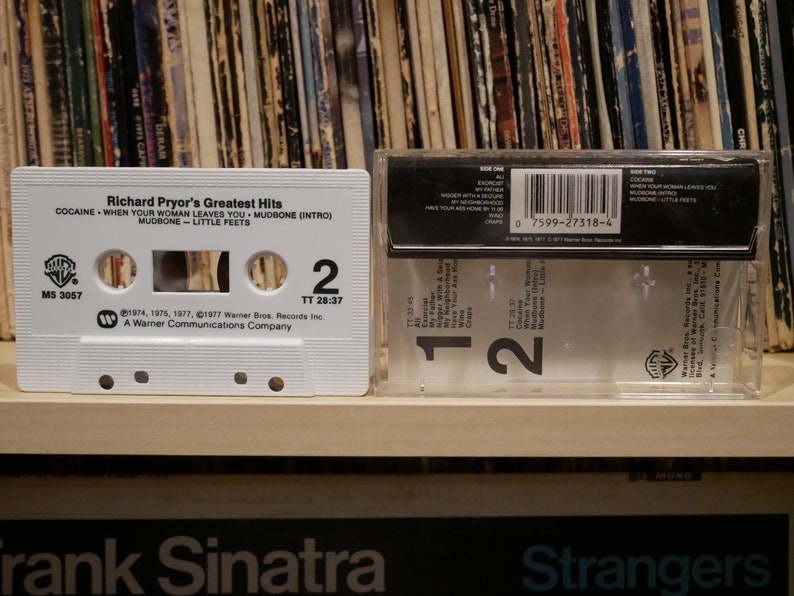 May include: A white cassette tape featuring "Richard Pryor's Greatest Hits" with song titles like "Cocaine" and "Mudbone." The cassette sits beside its clear plastic case, with a backdrop of vinyl records.