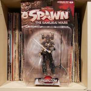 Può includere: Una action figure di Spawn di McFarlane Toys. La figura è una guerriera con un'armatura nera e un elmo da samurai. La figura tiene una spada e una lancia. La confezione è rossa e nera con le parole "Dark Ages Spawn The Samurai Wars" su di essa.