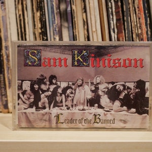 May include: A cassette tape with the album art for "Sam Kinison: Leader of the Banned." The artwork is a parody of The Last Supper, with Kinison as the central figure. The title and artist's name are in red gothic lettering.