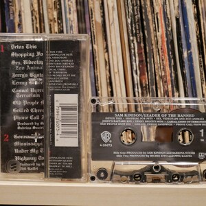 May include: A clear cassette tape of Sam Kinison's "Leader of the Banned" album. The cassette case displays the album title and track list, including "Detox This" and "Shopping for Pets." The background shows a collection of vinyl records.