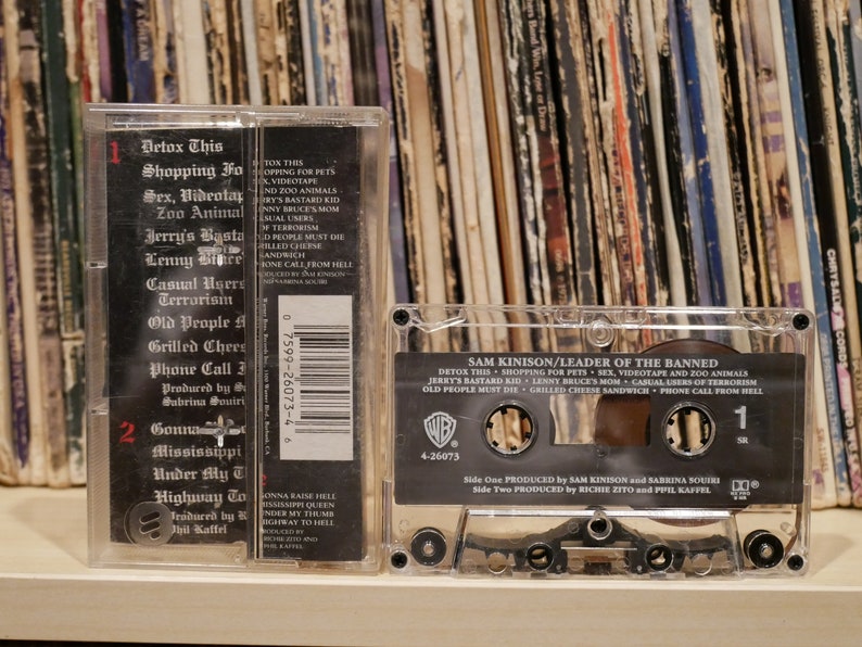 May include: A clear cassette tape of Sam Kinison's "Leader of the Banned" album. The cassette case displays the album title and track list, including "Detox This" and "Shopping for Pets." The background shows a collection of vinyl records.