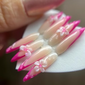 May include: Artificial nail set featuring a light pink base, vibrant pink tips, and delicate white flower embellishments. The nails are long and pointed, with a glossy finish. Small, clear droplets are also present.