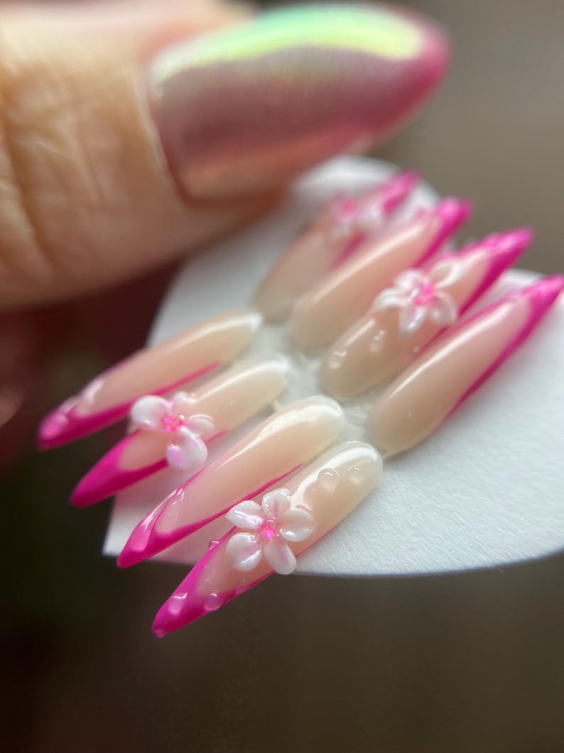 May include: Artificial nail set featuring a light pink base, vibrant pink tips, and delicate white flower embellishments. The nails are long and pointed, with a glossy finish. Small, clear droplets are also present.