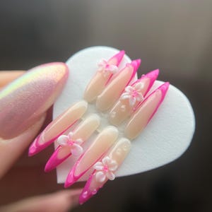 May include: A set of artificial nails with a French manicure design. The nails are long, almond-shaped, and feature a bright pink tip. Small, white, flower-shaped embellishments are placed on some of the nails.