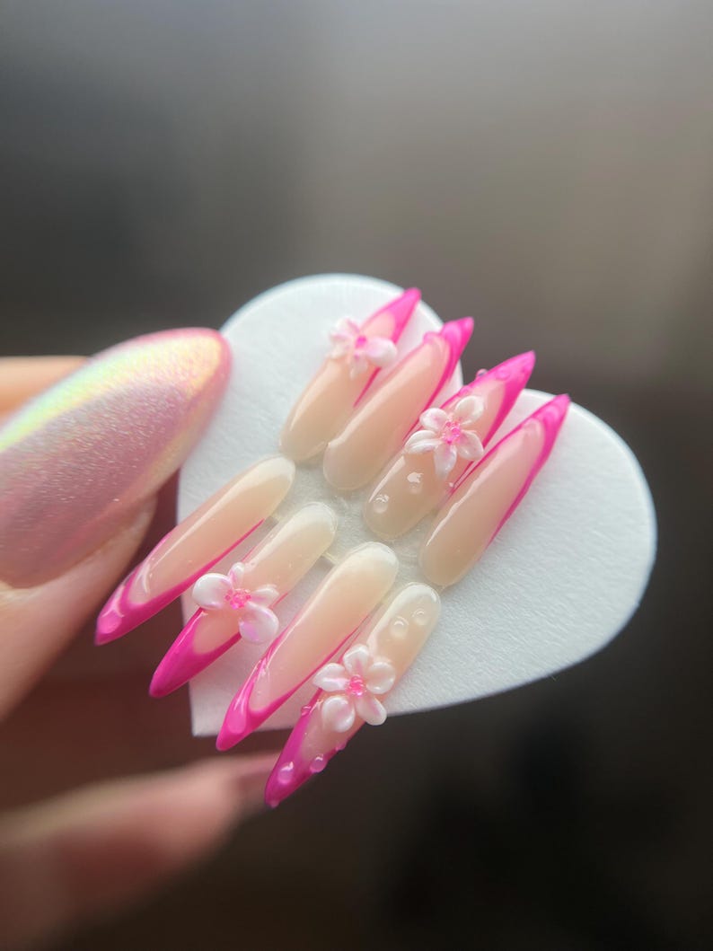 May include: A set of artificial nails with a French manicure design. The nails are long, almond-shaped, and feature a bright pink tip. Small, white, flower-shaped embellishments are placed on some of the nails.