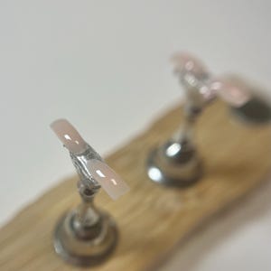 May include: A light pink and white French tip nail on a silver stand, displayed on a wooden surface. Another nail is visible in the background. The nails are a classic style.