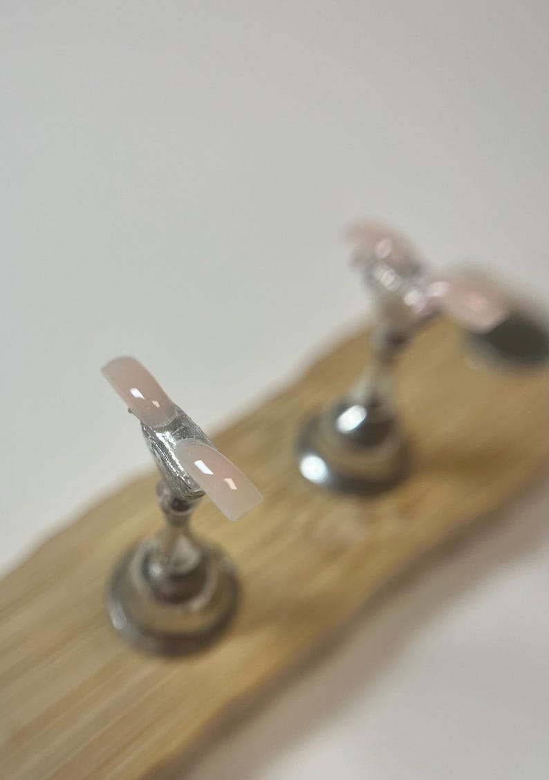 May include: A light pink and white French tip nail on a silver stand, displayed on a wooden surface. Another nail is visible in the background. The nails are a classic style.