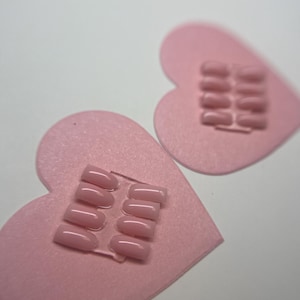 May include: Two sets of light pink press-on nails are displayed on pink heart-shaped felt cutouts. Each set includes several nails in a rounded square shape. The background is a plain white surface.