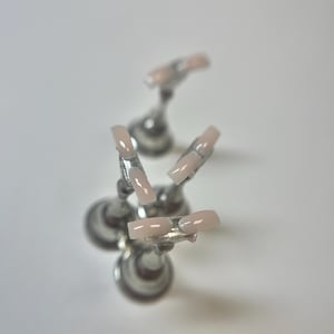 May include: Five light pink acrylic nails are displayed on silver stands. The square-shaped nails have a glossy finish. The stands are arranged in a circular pattern against a white background.
