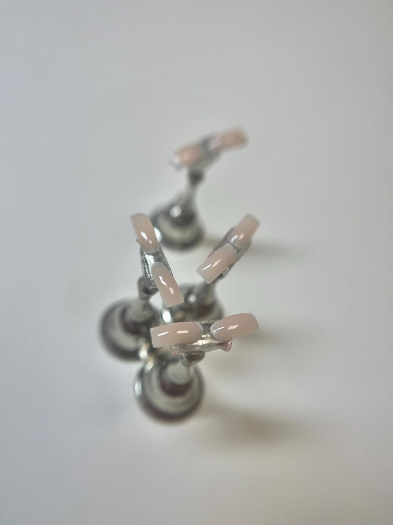 May include: Five light pink acrylic nails are displayed on silver stands. The square-shaped nails have a glossy finish. The stands are arranged in a circular pattern against a white background.
