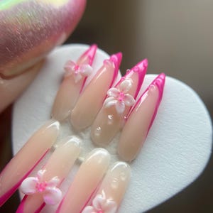 May include: A set of artificial nails with a long, almond shape. The nails have a light pink base with a bright pink French tip. Some nails are decorated with 3D white and pink flower embellishments. The nails are displayed on a white heart-shaped surface.