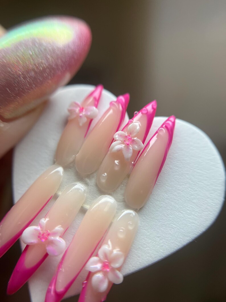 May include: A set of artificial nails with a long, almond shape. The nails have a light pink base with a bright pink French tip. Some nails are decorated with 3D white and pink flower embellishments. The nails are displayed on a white heart-shaped surface.