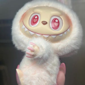 May include: A soft, white toy creature with a smiling face, red eyes, and sharp teeth. It has a furry hood with bunny ears and small hands with pink nails. The toy is a light pink colour.