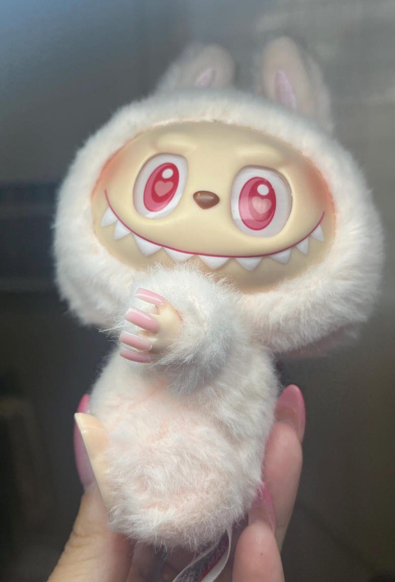 May include: A soft, white toy creature with a smiling face, red eyes, and sharp teeth. It has a furry hood with bunny ears and small hands with pink nails. The toy is a light pink colour.