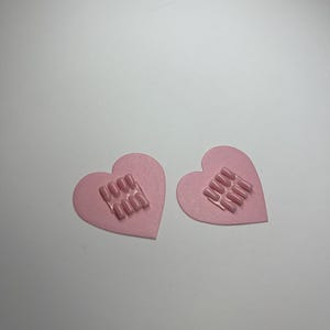 May include: Two pink heart-shaped nail art designs. Each heart features a rectangular arrangement of pink nail tips. The hearts are on a white surface. The nail art is a light pink colour.