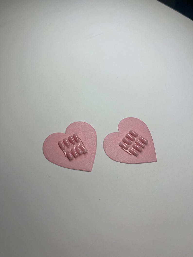 May include: Two pink heart-shaped nail art designs. Each heart features a rectangular arrangement of pink nail tips. The hearts are on a white surface. The nail art is a light pink colour.