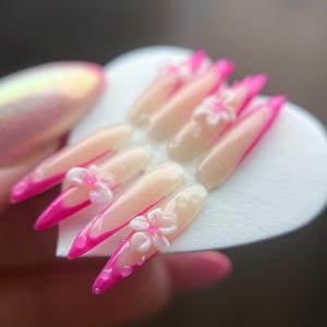 May include: A collection of long, pointed artificial nails. Each nail has a pale pink base, a vibrant pink tip, and a 3D white flower decoration. The nails are displayed on a white heart-shaped surface.